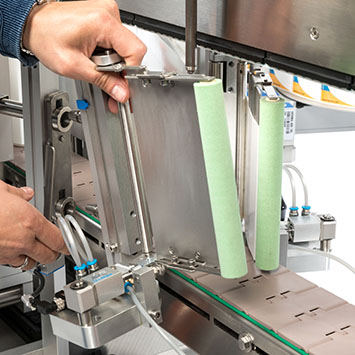 Eine Hand justiert eine Metallplatte an einer Maschine, während zwei grüne Rollen auf der rechten Seite sichtbar sind.