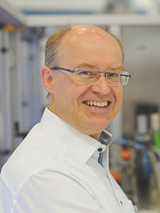 Ein Mann mit Brille und lächelndem Gesicht steht in einem Laborumfeld, umgeben von technischen Geräten und Ausrüstung.