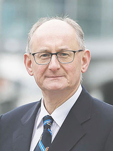 Ein Mann mit Brille und Anzug steht im Freien. Er hat graue Haare und trägt eine Krawatte mit Streifen.