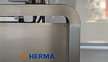 Ein Teil einer Maschine mit dem Logo "HERMA" in blauer Schrift auf einer metallischen Oberfläche.