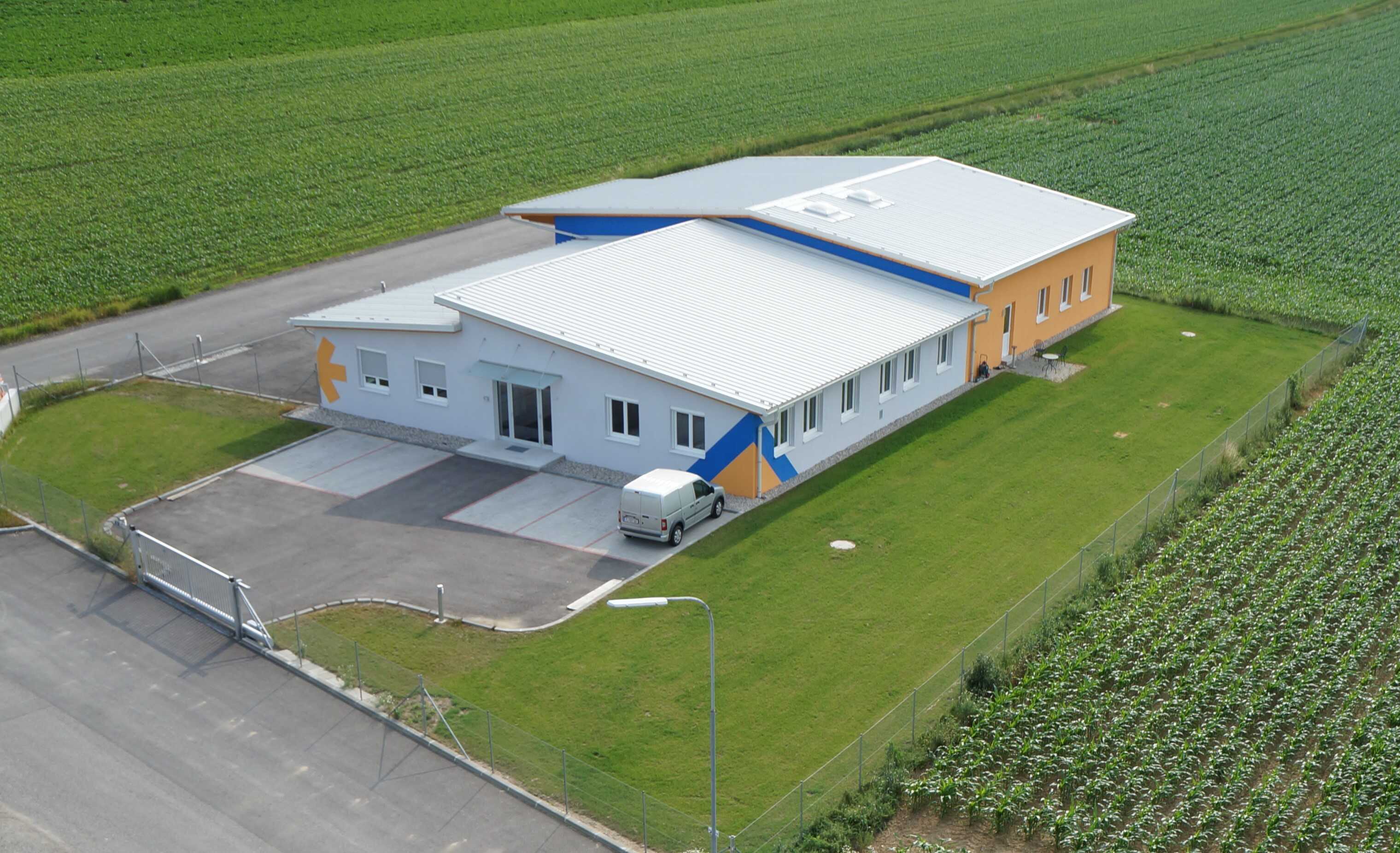 Ein modernes Gebäude mit einem flachen Dach, einer hellen Fassade und bunten Akzenten, umgeben von landwirtschaftlichen Feldern.