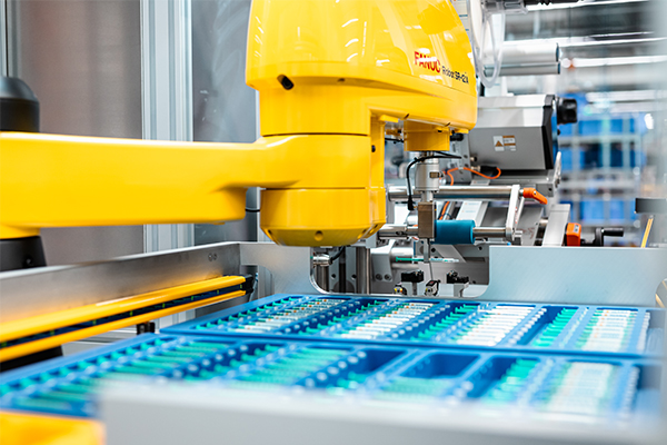 Ein gelber Industrieroboterarm arbeitet an einem Förderband mit blauen Behältern, die mit verschiedenen Objekten gefüllt sind.