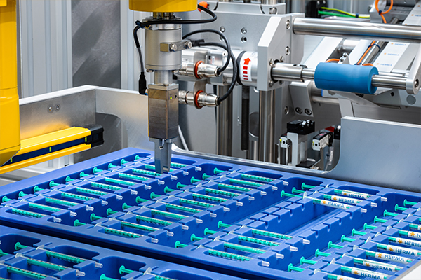 Automatisierte Maschine, die Spritzen in blauen Behältern bearbeitet, mit einem Roboterarm, der eine Spritze aufnimmt.