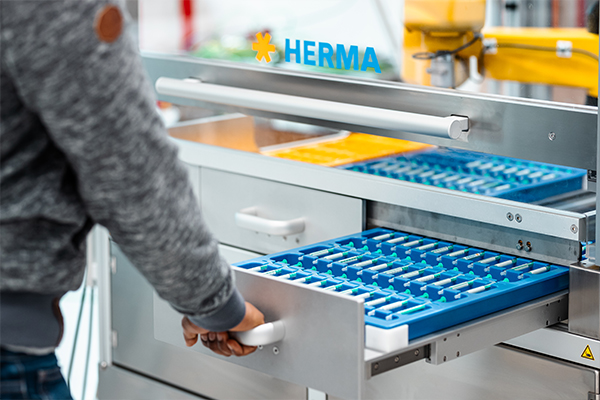 Eine Person zieht eine Schublade mit blauen Fächern aus einem Regal, das mit dem Logo "HERMA" gekennzeichnet ist.