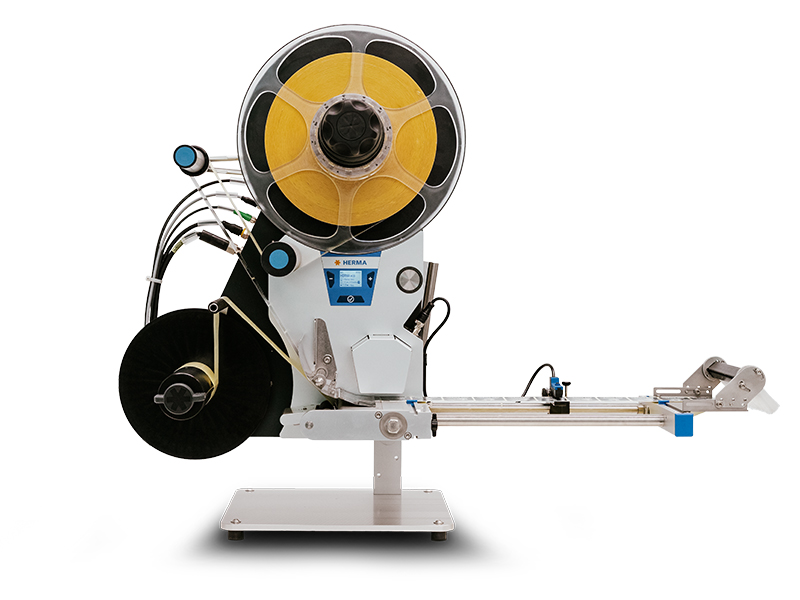 Eine Etikettiermaschine mit einer großen Spule, die gelbe Etiketten auf einem Förderband anbringt. Sie hat mehrere Anschlüsse und Bedienelemente.