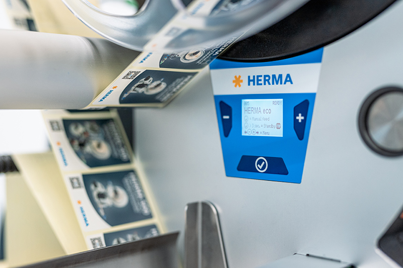 Ein Etikettendrucker mit einem Display, das die Marke HERMA zeigt. Etiketten werden auf einer Rolle ausgegeben.