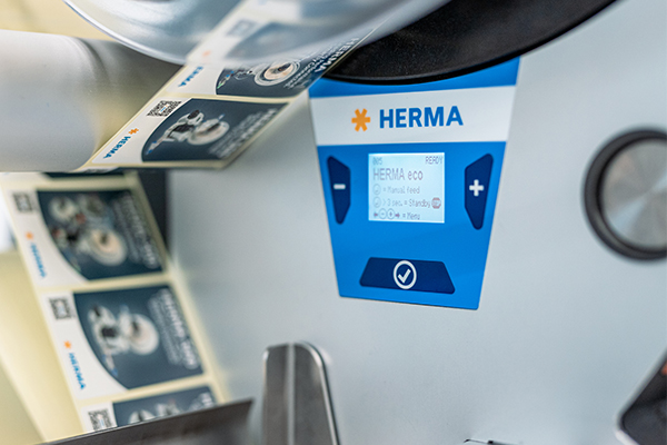 Ein Etikettendrucker mit einem digitalen Bedienfeld, das die Marke "HERMA" zeigt. Etikettenrollen sind sichtbar.