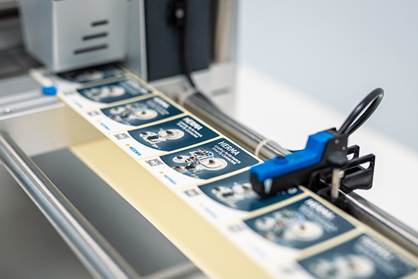 Eine Druckmaschine verarbeitet Etiketten mit technischen Illustrationen und Text. Die Etiketten sind auf einer Rolle angeordnet.