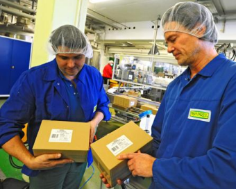 Zwei Arbeiter in blauen Overalls und Haarnetzen halten Kartons in einer Produktionsumgebung und überprüfen deren Inhalte.
