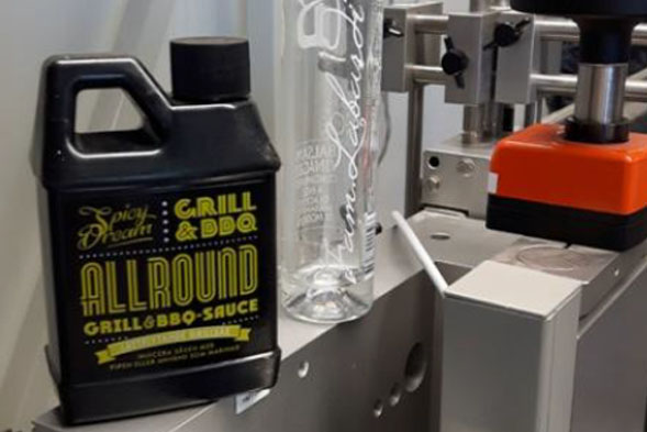 Eine Flasche Grillsoße und ein transparentes Glas stehen auf einer Maschine in einer Produktionsumgebung.