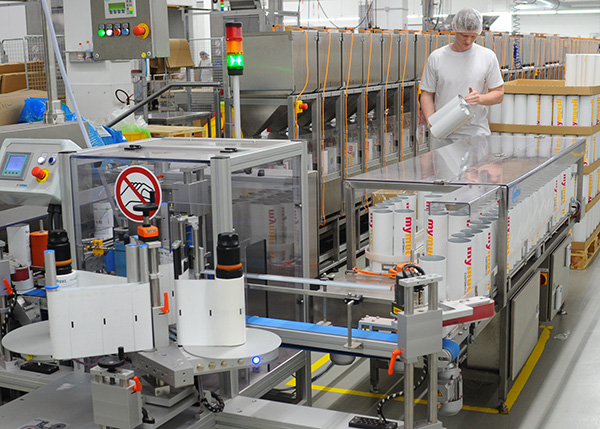 Ein Arbeiter in einem Produktionsbetrieb füllt Behälter an einer automatisierten Abfülllinie. Maschinen und Verpackungen sind im Hintergrund sichtbar.