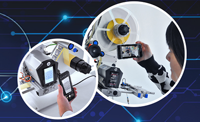 Eine Person hält ein Smartphone, während sie eine Maschine mit einem Bildschirm und einem anderen Gerät betrachtet. Technologische Elemente sind im Hintergrund sichtbar.