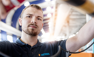 Ein Mann in einem HERMA-Polohemd arbeitet an elektrischen Kabeln in einer modernen Umgebung mit hellem Licht.