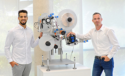 Zwei Männer in weißen Hemden stehen neben einer technischen Maschine mit Rollen und einem Display, die für industrielle Anwendungen konzipiert ist.