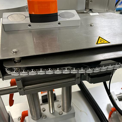 Eine Maschinenkomponente mit einem orangefarbenen Element, Metalloberflächen und einem Kettenantrieb, die in einer industriellen Umgebung montiert ist.