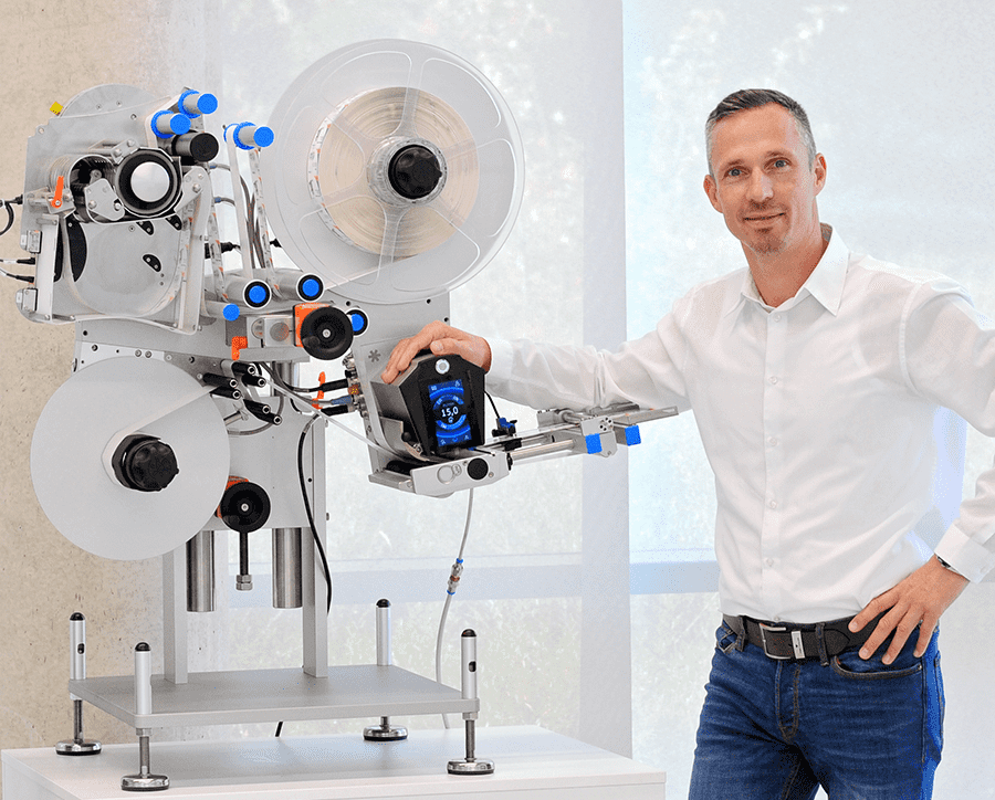 Ein Mann steht neben einer modernen Etikettiermaschine und hält ein Bediengerät in der Hand. Die Maschine hat mehrere Rollen und Steuerungselemente.