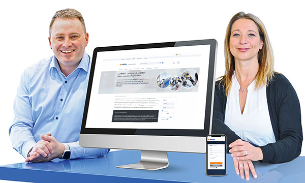 Zwei Personen sitzen an einem Tisch, neben einem Computer und einem Smartphone, die eine Webseite anzeigen.