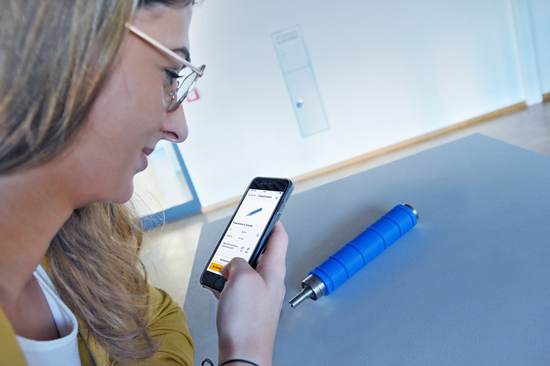Eine Person mit Brille schaut auf ihr Smartphone, während sie einen blauen Zylinder auf einem Tisch betrachtet.