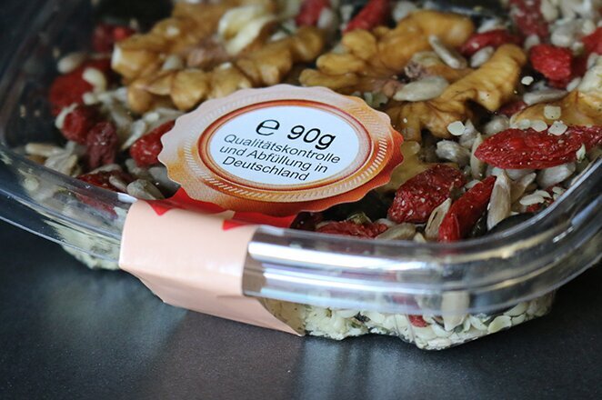 Eine transparente Schale mit einer Mischung aus Nüssen, Samen und getrockneten Früchten, etikettiert mit Nährwertinformationen.
