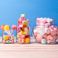 Bunte Süßigkeiten in Gläsern, darunter Lutscher, Fruchtgummis und Herz-Marschmallows, auf einem hellen Holzuntergrund.