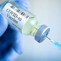 Eine Hand mit blauen Handschuhen hält eine COVID-19-Impfstoffflasche, während eine Spritze bereit ist, um die Flüssigkeit zu entnehmen.