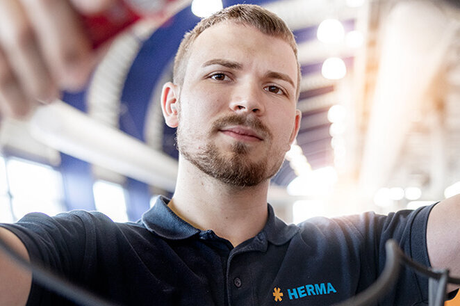 Ein junger Mann in Arbeitskleidung hält Kabel in einer modernen Umgebung mit einem Gebäude und Bäumen im Hintergrund.