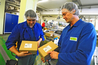 Zwei Männer in blauen Arbeitsanzügen und Haarnetzen halten Kartons in einer Produktionsumgebung und überprüfen deren Inhalte.
